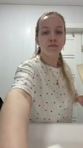 A young woman in a white shirt with a brown heart pattern stands in a bathroom in front of a white sink and a white door.  She opens a white tube with one hand and squeezes it, dispensing white cream or lotion onto her opposite palm.