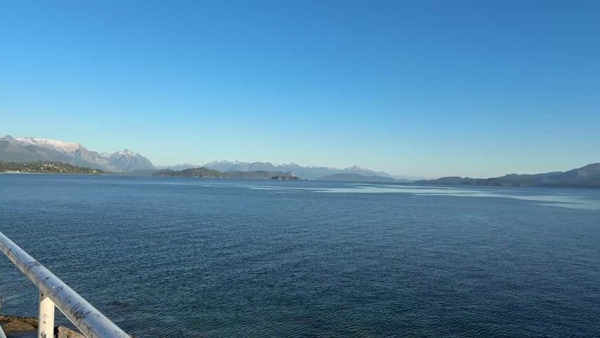 The camera pans across a body of water with a clear, blue sky above. A mountain range is visible in the distance. Some of the mountain peaks are snow-capped. The water is calm with gentle ripples. A portion of a white railing is visible in the bottom left corner of the frame, suggesting the view is from a boat or a pier. The scene is bright and well-lit.