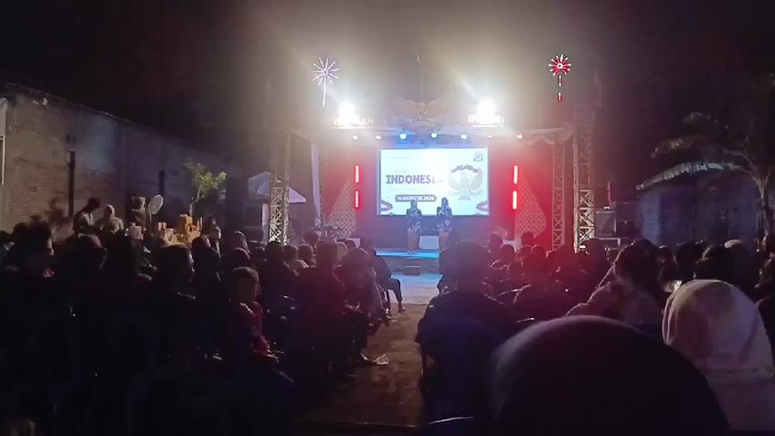The video shows an outdoor night-time event with a stage and an audience. On the stage, there is a large screen displaying the word "INDONESIA" and what appears to be an emblem or logo. Two people are standing on the stage in front of the screen, seemingly addressing the audience. The stage is lit with bright lights, including some decorative festive lights at the top. Red and white banners are also visible behind the screen. The audience is seated in chairs facing the stage, with many people visible in the foreground, middle ground, and background. The environment appears to be somewhat rustic, with brick walls and some vegetation visible on the sides. The overall atmosphere suggests a cultural or public gathering.