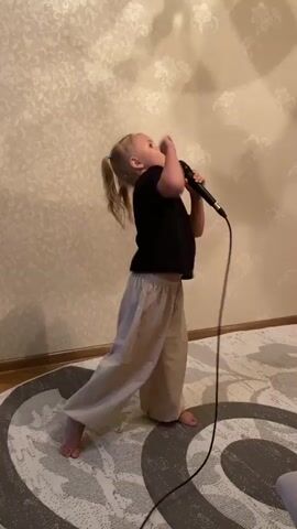 A young girl with blonde pigtails stands on a rug in front of a patterned wall, holding a microphone and singing. She is wearing a black t-shirt and loose, tan pants. Her right arm is raised as if gesturing while she sings into the microphone held in her right hand. The microphone is connected to a wire that trails on the floor.