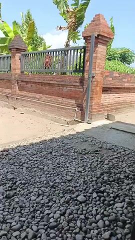 The video shows a fence made of brick and metal, with decorative elements on the fence. Behind the fence are banana trees and other greenery. The ground in front of the fence is covered with gravel and sand. There are also two concrete structures that appear to be bases for something, possibly statues or lamps. A pair of flip-flops is visible on the sandy ground. The sky is blue with some clouds, indicating it is daytime and sunny.