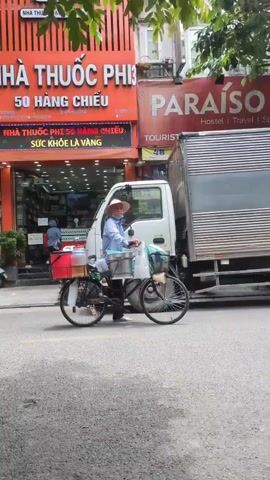 A street scene unfolds with a woman in a straw hat on a bicycle laden with containers and bags. She is positioned between a white delivery truck and a large silver truck with visible license plates. To the left, a motorcycle with two passengers, a woman wearing a green helmet and a blue top, and another person, are visible. Further down the street, another motorcycle is seen with two individuals wearing hoods. Businesses with signage in Vietnamese and English are visible in the background, including "NHA THUOC PHI" and "PARAISO Hostel Travel Spa." The environment appears to be a busy urban street with trees and buildings.