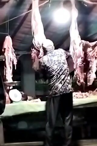 A man wearing a printed shirt and a light-colored cap stands behind a table, working with meat. Several large pieces of meat hang from above. A scale is visible on the table. A second person stands to the left, wearing a dark jacket. A single bright light illuminates the scene. The environment appears to be a butcher shop or market stall.