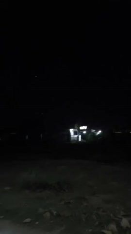 The video shows a dark, outdoor scene at night. A dirt or gravel road is visible in the foreground, with vegetation on the sides. In the distance, there are lights from buildings and possibly streetlights. The sky is dark with a few faint points of light, which could be stars or distant lights. The camera appears to be moving forward along the road. There are no people or animals visible in the video. The overall impression is one of a quiet, rural or semi-rural area at night.