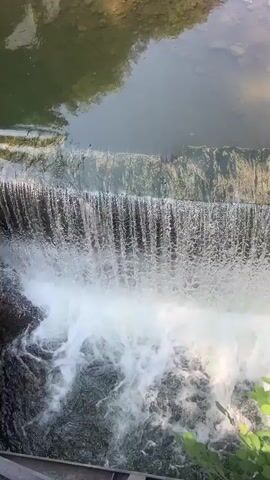 The video shows a close-up, top-down view of a small waterfall cascading into a pool of water below. The water at the top of the waterfall is relatively still and appears green, reflecting the surrounding foliage. As the water goes over the edge, it forms a curtain of white, frothy water. The pool below is turbulent, with white foam and swirling water. The bottom of the pool is rocky and appears shallow in places. Along the edge of the upper pool, there is a man-made structure made of concrete or stone. Further upstream, a wooden boat-like structure is visible, resting on a ramp leading into the water. The water appears to be clear enough to see the bottom in some areas. The overall scene is one of a natural water feature within a constructed environment.