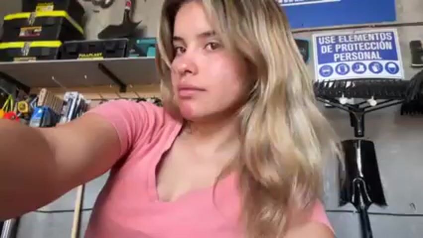The video shows a woman with long blonde hair and a pink shirt standing in what appears to be a garage or workshop. She has a red mark on her face near her jawline. The background includes tools and equipment such as toolboxes, shovels, rakes, and various hand tools hanging on a wall. A sign is visible in the background with the words "USE ELEMENTOS DE PROTECCIÓN PERSONAL" along with several icons of safety equipment. The woman looks at the camera and holds it at arm's length, suggesting she is taking a selfie. There is audio playing in the background.