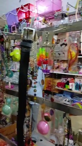 The video shows an overstuffed shop with a variety of colorful items, mainly jewelry.  The camera pans across shelves and racks displaying necklaces, bracelets, earrings, and other accessories. The items are colorful, and mostly made of plastic or beads.  Some of the shelves also hold packaged goods and other miscellaneous items. A person can be seen briefly standing in front of the rack of items.  The shop appears small and cluttered.