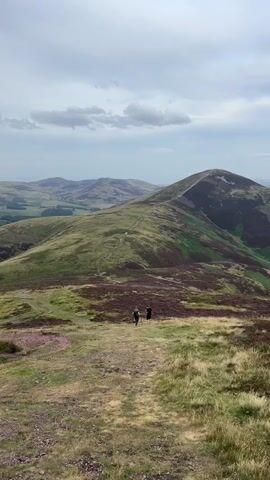 The video shows a wide shot of a rolling mountain landscape under a cloudy sky. In the foreground, a grassy path winds down a hill. Two people, appearing to be hikers, are walking on the path in the middle ground. The hills are covered in green grass and patches of purple heather. In the background, more rolling hills stretch into the distance. The lighting suggests it is daytime, possibly late afternoon.