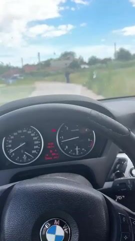 The video shows a first-person perspective from inside a car, looking forward through the windshield. The car's dashboard is visible, displaying a speedometer, tachometer, and digital readouts including speed, engine temperature, and trip mileage. The road ahead is unpaved, with dry grass and some vegetation on either side. In the distance, a person dressed in dark clothing, possibly a hooded jacket, is walking away from the car. This person appears to be using a walking stick. Several buildings and trees are visible in the background, with a cloudy blue sky overhead. The car's steering wheel is also in the foreground. The timestamp on the dashboard indicates the time as 13:22.