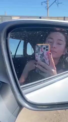 The video shows a woman taking a selfie in the side mirror of a white car. The mirror is splattered with dirt and water spots. The woman has dark hair, dark nail polish, and is wearing a black top. She is holding a cell phone with a floral case. Outside the car, a freight train is passing by on a track parallel to the road. The sky is blue, and utility poles are visible in the background. The environment appears to be somewhat rural or industrial.