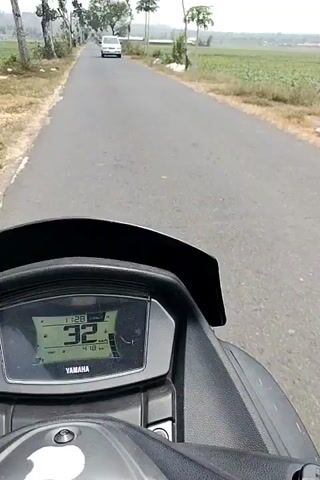 The video shows a first-person perspective from a motorcycle or scooter. The rider is on a paved road with trees lining the sides and fields in the background. A white minivan is visible on the road ahead. The digital dashboard of the vehicle shows a time of 11:28, a speed of 32 km/h, and a trip distance of 4.19 km. The speed increases to 33 km/h and then 35 km/h as the video progresses. The environment appears to be rural and daytime.