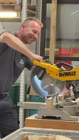 A middle-aged man with short blonde hair, wearing a dark grey polo shirt and dark grey work pants, is standing in a workshop. He is smiling and operating a yellow and black DeWalt brand miter saw. The saw blade is spinning and cutting a piece of light-colored wood that is secured to the saw's base. The man is holding the saw's handle with his right hand. In the background, there are shelves with various items, including what appears to be wood, boxes, and some lighting fixtures. The environment suggests a woodworking or construction setting.