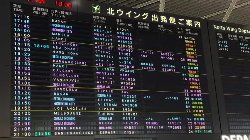 The video shows a close-up of a large, illuminated departure board at an airport. The board displays flight information in both Japanese and English. The primary language is Japanese, with headings like "航空会社" (Airline), "便名" (Flight Number), "行き先/経由地" (Destination/Via), and "ゲート" (Gate). The English section, labeled "North Wing Departure," contains similar information. The board is segmented into columns, listing flight times, airline names, flight numbers, destinations, gate numbers, and remarks. The times displayed range from 17:10 to 21:55. Several flights are listed with various destinations including Busan, Ho Chi Minh City, Manila, Hanoi, Singapore, Abu Dhabi, Hong Kong, Seoul, Bangkok, Calgary, Bengaluru, Vancouver, Honolulu, Los Angeles, Melbourne, and San Francisco. Some entries have specific gate numbers, while others indicate "GO TO GATE" or "GATE CLOSE." The lighting is dim, suggesting it might be evening or indoors. The screen is digital and appears to be constantly updating or displaying information. There are no people visible in the frame, only the information display itself. The overall impression is one of a busy, functional airport environment focused on providing flight departure information.