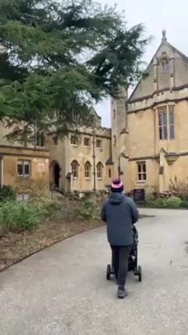 A person wearing a winter hat and blue jacket is walking away from the camera pushing a stroller on a paved pathway. The pathway curves gently to the left. The person and stroller are walking towards a multi-story stone building.  A large tree stands to the left of the pathway, and shrubbery and plants are on either side of the path. The sky appears to be overcast.
