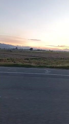 A video taken from a moving vehicle shows a road with a white line. To the right of the road are fields with some crops. The sky is clear with a few clouds and the sun is setting, casting a warm glow on the horizon. In the distance, there are some trees and buildings. On the right side of the road, there is a truck, a motorcycle, and a few people. The video pans to the right, showing more of the fields and the sunset.