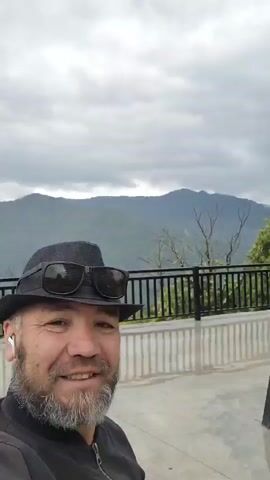 A man in a black fedora and sunglasses is taking a selfie. He has a gray beard and is wearing a black jacket. In the background, there are mountains under a cloudy sky. The man is smiling and looking at the camera.