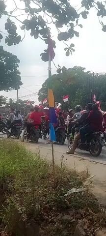 The video shows a street scene with a procession of motorcycles. The motorcycles are predominantly ridden by people wearing red and white clothing. Many of the motorcycles are decorated with Indonesian flags, which are red and white. In the foreground, there is a tall pole with colorful fabric streamers attached to it, consisting of blue, purple, yellow, and red sections. The motorcycles are moving from left to right in the frame, with some people standing on the side of the road watching. The environment appears to be outdoors, with trees and vegetation visible in the background. The sky is overcast. The overall impression is of a celebratory event or parade.