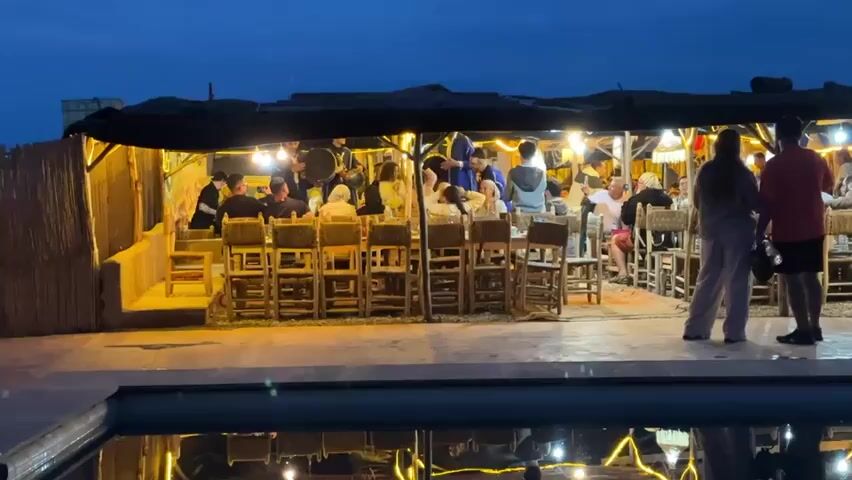 The video captures an outdoor evening gathering in Lalla Takarkoust, Morocco. A group of people, including adults and children, are seated at long, rustic wooden tables with matching chairs, enjoying a meal. The dining area is set under a dark canopy or tent-like structure and is brightly illuminated by numerous string lights, casting a warm glow.

In the center of the dining area, several men dressed in traditional attire are performing live music. They are playing large, round drums and possibly other instruments, creating a lively and festive atmosphere. Some diners are actively watching the performance, while others appear to be conversing or recording the event with their mobile phones.

In the foreground, a swimming pool reflects the warm lights of the dining area, enhancing the ambiance. The sky is dark blue, indicating it is nighttime. The overall scene suggests a cultural dining experience with traditional Moroccan entertainment. There is no visible text.