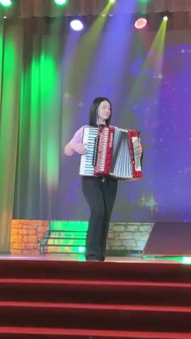 A young woman stands on a red-carpeted stage playing an accordion. She is centered on the frame, wearing a patterned short-sleeved shirt and black pants. The accordion is primarily red and white with decorative details. Behind her, a vibrant backdrop projects stars and sparkles against a purple background. Green stage lights illuminate the left side of the stage, and other lights shine from above. A faux stone platform is visible behind her, and she stands on a tiered platform. The camera remains static throughout the video.