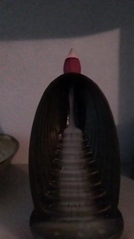 A still image shows a dark brown ceramic object, possibly a vase or decorative piece, with a red and white cone-shaped object on top. The ceramic piece has an egg-like shape with vertical striations and a lighter interior that suggests a tunnel or pathway. It sits on a light-colored surface, presumably a shelf or table, against a pale blue or gray wall. There are other objects, slightly out of focus, on either side of the ceramic piece. The lighting appears to be dim.