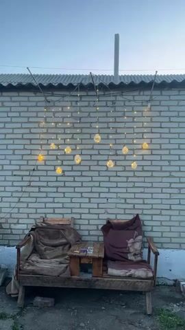 A vertical video shows two chairs and a small table in front of a brick wall decorated with string lights. The chairs are worn and covered with blankets. The table has a cigarette pack and a glass on it. The lights are lit, creating a warm glow. The sky above is a pale blue, indicating it is dusk or dawn.