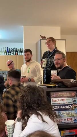 Here is a detailed description of the video:

A group of approximately ten people are gathered indoors, seemingly in a living room or common area. The camera captures the scene from a slightly low angle, showing the group clustered together, with some individuals standing and others seated.

The focal point is a woman with glasses and a ponytail. She is standing on a slightly elevated position and appears to be speaking to the group. She is holding a cup in her hand. To her left, there are two men standing, one with a watch on his left wrist and a green object clipped to his shirt, and the other with glasses and a beard.

In the background, a man with an eye patch stands among other people. To the right of the woman, there's a black shelf holding stacked board games like "Ticket to Ride," "Small World," and "Dominion". Above the shelf and behind the woman, there is a white kitchen cabinet on which there's a small brown dachshund figurine. To the left, a rack of wine bottles is visible.

People are holding cups and appear to be listening attentively. The overall atmosphere seems informal and conversational.