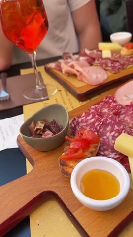 The video shows an outdoor table with two people seated. On the table are two wooden boards of assorted meats and cheeses, a small bowl of what appears to be dried fruit, a bowl of honey, and small toasted bread pieces with tomatoes. Two glasses of red-orange cocktails with ice are also visible. Additionally, two wooden boxes containing fried bread or dough are present. A fork, a Fiat car key with an Italian flag keychain, and a napkin are also on the table. One person is wearing a white t-shirt, and the other is wearing a bright pink shirt. The setting appears to be a restaurant or cafe, as evidenced by the table setting and background noise of conversations.