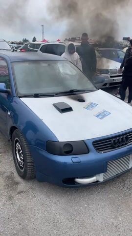 A blue Audi A3 is parked outside, with a white hood that has a large vent. The hood also has soot marks and event stickers on it. The front bumper is modified with an intercooler installed. People are gathered around the car, and black smoke is billowing from the vent on the hood. A popping noise is heard repeatedly. The setting appears to be a gathering of car enthusiasts.