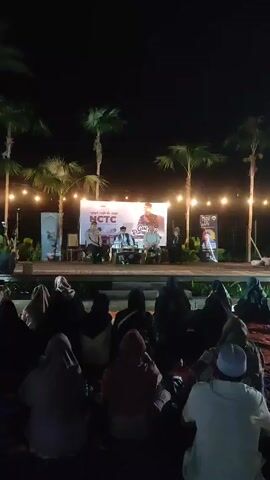 Here is a description of the video:

The video shows an outdoor nighttime event. An audience is seated on the ground, facing a stage set up with a backdrop and several people seated at a table.  The backdrop has text and a picture of a person on it. Palm trees surround the stage, and string lights are hung around the area. It seems to be a gathering or event, possibly a talk or lecture, taking place at night.