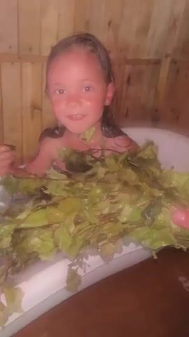 A young girl with dark hair is in a bathtub. She is holding a bundle of green leaves. The bathtub is in a room with wooden walls. The girl smiles at the camera. She splashes the leaves in the water.