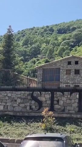 This is a vertical video clip filmed on a sunny day in Setcases, Spain. The scene opens with a view of a stone building, likely a house or inn, set against a backdrop of a lush, green mountainside covered in trees. The building has a rustic appearance with stone walls and a tiled roof. A metal fence runs in front of the building, and large, dark letters spelling out "SETO" are visible on a stone wall below the fence.

As the video progresses, the camera pans to the right, revealing more of the stone building and its surroundings. We see additional windows and architectural details on the building, and the mountainous terrain continues to dominate the background. The sun is clearly visible, casting bright light and creating shadows, indicating it's likely mid-day. The sky is a clear, vibrant blue, suggesting fair weather.

Further to the right, the camera captures another section of the stone building, and then a sign is visible on an upper section of the building that reads "Ca La Núria." This suggests the building is likely an establishment named "Ca La Núria." The clip ends with a view of more of the stone architecture and the continued green mountainous landscape. Cars are visible in the foreground at the bottom of the frame, suggesting this is a road or parking area near the building. The overall impression is one of a picturesque, rural mountain village in Spain.