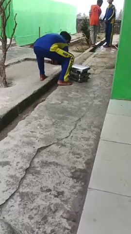 The video shows a young person in a blue and yellow tracksuit picking up a stack of what appear to be metal trays. They are barefoot and carrying the trays with their right arm. In the background, two other young people are visible, one in an orange shirt and the other in a blue and yellow tracksuit. They are standing near a green wall and some foliage. The ground is paved with concrete. The scene appears to be an outdoor area, possibly a schoolyard or a communal dining area.