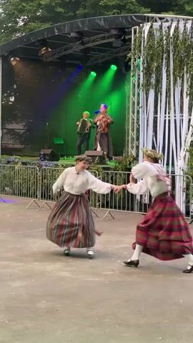 Two young women in traditional folk costumes, featuring long plaid skirts and white blouses, dance in front of a raised outdoor stage. They are facing each other and appear to be performing a choreographed dance. The woman on the right wears a red and green plaid skirt and a white blouse with red embroidery, her hair is styled in braids adorned with flowers. The woman on the left wears a striped skirt with red, white, and gray stripes and a similar white blouse, her hair also adorned with flowers. Both women are barefoot.

On the stage, three musicians are performing. In the center, a person in a dark blue cloak and a purple headdress plays the violin. To the left of the violinist, a person in a dark suit plays an accordion. To the right of the violinist, another person stands, possibly singing or playing an instrument, wearing a dark cloak and a headdress. Green and blue stage lights illuminate the performers from behind.

The background shows a black stage curtain and the metal structure of the stage rigging. To the right of the stage, tall, decorative poles draped with white ribbons and green foliage create an entrance or frame. A metal fence with green mesh separates the performance area from the open space where the dancers are. The ground is paved with a rough, gray material. Trees are visible in the upper background, suggesting an outdoor evening event.