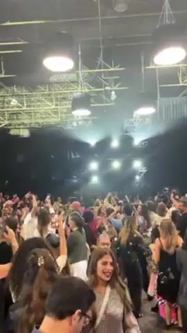 A crowded event space features numerous people, illuminated by bright overhead lights. The crowd appears to be a mix of ages and genders, many recording with their phones. The atmosphere seems lively with a stage visible in the distance, shrouded in light. The space has an industrial feel, with exposed ceiling beams and lighting rigs.