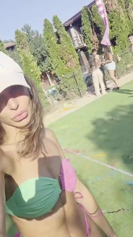 A woman in a green and pink bikini takes a selfie with a blurry background. She is outdoors, with grass and trees visible. Other people, out of focus, are in the background. One person is wearing a white baseball cap and appears to be smiling. The woman is showing her midriff and a portion of her legs. The video seems to be at a daytime event, possibly a pool party or festival, with a pink banner visible in the background.