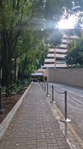 The video shows a paved pedestrian walkway next to a road. Trees line the walkway on the left, and a large building, possibly a parking garage, is visible on the right. A chain barrier separates the walkway from the road. In the distance, several people are walking along the path. The sun is shining, casting light and shadows across the scene.