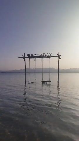 The video displays a serene scene featuring a wooden swing set standing in calm, clear water. The swing set has two swings, both partially submerged and gently swaying. Above the swings, the word "SOMABAY" is prominently displayed in dark letters on the top beam of the structure.

In the background, a distant mountain range can be seen along the horizon, silhouetted against the sky. The sky itself transitions from a soft blue-purple on the left to a lighter, almost white or pale yellow on the right, suggesting either early morning or late afternoon light. The sun, though not directly visible, seems to be low on the horizon, casting a gentle glow. The water reflects the light and the structures, creating a peaceful and picturesque ambiance. No people are visible in the shot. The overall impression is one of tranquility and natural beauty, characteristic of a resort or beach destination in Ras Abu Soma, Egypt.