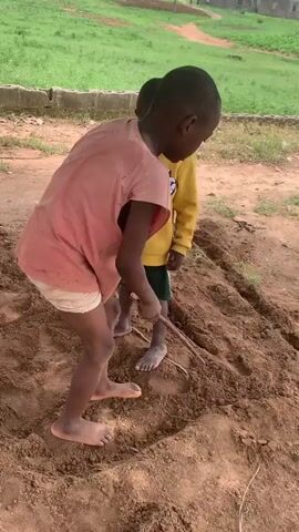 Two young boys are playing in the dirt outdoors. One boy, wearing a pink shirt and light shorts, uses a stick to carve a large circle in the dirt. The other boy, wearing a yellow sweatshirt and green shorts, stands nearby and watches. They are both barefoot. The area around them is dirt and grass. There are some concrete structures in the background.