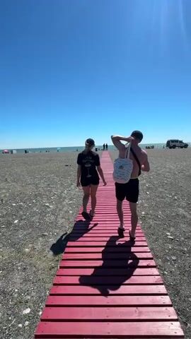 A man and a woman walk hand-in-hand down a red wooden boardwalk on a beach. The woman is on the left, wearing a black t-shirt and shorts. The man is on the right, wearing black shorts and carrying a white bag over his shoulder. They are walking away from the camera towards the ocean, which is visible in the distance. The sky is clear and blue, and the beach is covered in pebbles. There are other people on the beach and in the water.