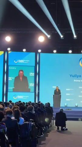 The media file shows a woman speaking at a podium during the Ukraine Recovery Conference in Gdańsk, Poland. The woman, likely a speaker at the event, is wearing a dark suit and glasses. She is standing behind a dark grey podium with a microphone. The backdrop is a large blue screen displaying the "UkraineRecovery Conference" logo. To the left of the main screen, there is another screen showing a close-up of the speaker. The scene is an indoor conference hall, with rows of chairs filled with attendees visible in the foreground. The lighting suggests it's daytime or the event is well-lit for a presentation. The atmosphere appears formal and professional, typical of a significant international conference. The visible text confirms the event as the "UkraineRecovery Conference" and identifies the speaker as "Yuliya" on the main screen.