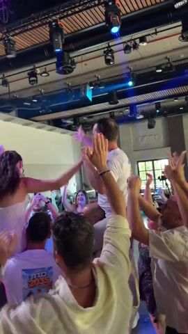 This video shows a lively wedding reception. A man and woman are seated on the shoulders of other men in the center of a dance floor, being lifted and cheered by a crowd. The man, wearing a white t-shirt, is looking forward and raising his arms. The woman, in a sparkly white dress, has her back to the camera and is also raising her arms. The surrounding guests are dressed in white and are clapping and dancing. The environment appears to be an indoor venue with a high ceiling, stage lighting, and exposed beams.