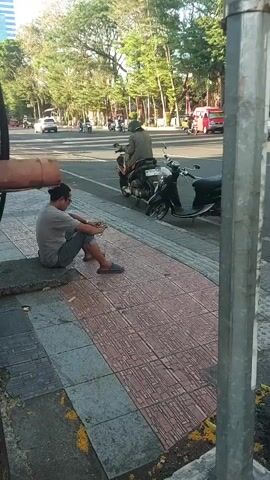 A person sits on a curb, looking down, with their hands clasped. They are wearing a gray t-shirt and dark pants. Behind them, a scooter is parked. Further back, a person on a motorcycle rides past, wearing a helmet and a green jacket. There are trees and other vehicles on a street in the background. The scene appears to be an outdoor public space, possibly a sidewalk next to a road.