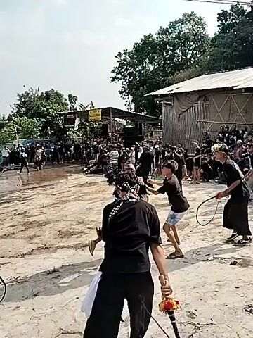 A crowd of people are gathered in an outdoor, dusty area. In the center, several young men are engaged in what appears to be a ritualistic or performative act. Two young men, dressed in black shirts and shorts, are facing each other and appear to be interacting physically, possibly in a form of sparring or dance. One of them is holding a whip. Another young man in a black shirt and pants with a bandana around his head is walking away from the center of the activity, carrying a white bag. Behind the central group, a larger crowd of people of various ages are watching the event. Some people are sitting on the ground, while others are standing. The background shows simple structures, possibly houses or stalls, with corrugated roofs and wooden walls, surrounded by trees. The sky is clear and bright. The overall atmosphere seems to be one of a community gathering or event.