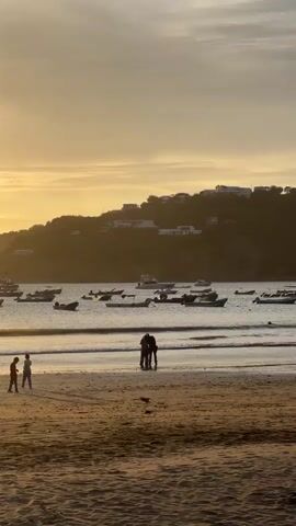 The video shows a beach scene at sunset. In the foreground, a sandy beach is visible with a few people. Further back, the ocean has gentle waves rolling onto the shore. Numerous small boats are anchored in the water, and a larger white boat is also present. In the background, a hill covered with vegetation and houses overlooks the bay. The sky is a soft orange and yellow, indicating the sun is setting. Two people are standing close together near the water's edge, appearing to be embracing or conversing. Another person is standing further to the left on the beach. The overall atmosphere is peaceful and serene.