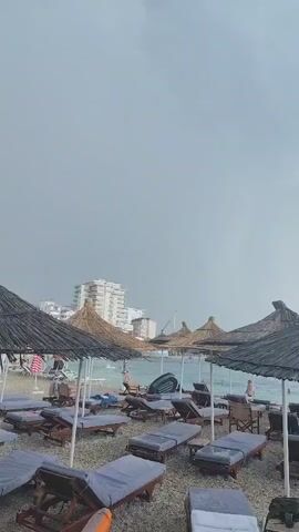 The video shows a beach with numerous lounge chairs and thatched umbrellas. The sky is overcast and appears to be either before a storm or during a storm, indicated by the dark, cloudy sky. In the background, buildings are visible along the coastline. Some people are present on the beach, some sitting on lounge chairs, and others appear to be in or near the water. There is an inflatable object, possibly a float, in the foreground of the last frame. The overall atmosphere is subdued due to the weather conditions.