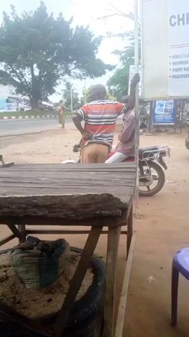 This media file captures an outdoor street scene in Mbribit, Nigeria, during the daytime, under what appears to be an overcast or hazy sky. In the foreground, a rustic wooden table stands, beneath which a sandbag rests on an old tire.

Two men are the primary subjects near the center of the frame. One man, wearing an orange and white striped shirt and light brown shorts, stands with his hands on his hips, facing away from the camera and looking towards the road. Beside him, another man in a lighter-colored shirt sits on a motorcycle, holding onto a white signpost.

The background features an asphalt road with a woman in a patterned dress walking on the opposite side. Other indistinct figures and buildings can be seen further in the distance, alongside mature trees. The white sign that the man is holding onto has visible text fragments, including "CH", "BIBLE CL", "MIDWE", "TARRY N", and "PASSOV", suggesting it is a church sign advertising various activities like "Bible Class," "Midweek," "Tarry Night," and "Passover." A partially visible purple plastic chair is also present on the right side of the foreground. The overall scene depicts a casual moment in a bustling local environment.