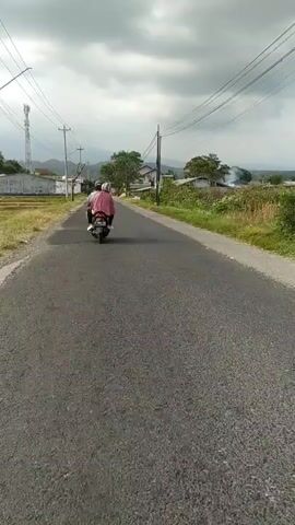 A person rides a motorcycle down a paved road. The motorcycle is traveling away from the camera. The road is lined with vegetation and houses. Power lines are visible overhead. The sky is cloudy.