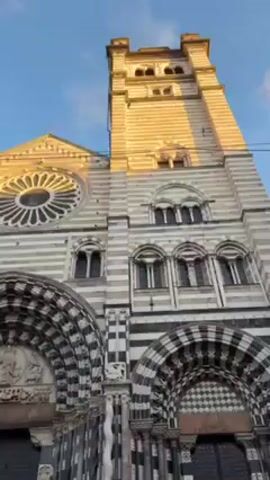 The video shows the Cathedral of San Lorenzo in Genoa, Italy, from a low-angle perspective. The cathedral has a facade made of alternating white and dark gray marble stripes, a large rose window, and a tall tower. People are standing and sitting on the steps in front of the cathedral's entrance. The sky is blue with a hint of golden light.