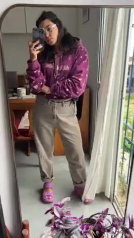 A woman wearing glasses is taking a selfie in front of a mirror. She is wearing a purple hoodie with a pattern, beige pants rolled at the ankles, and bright pink sandals. She is holding a black smartphone. The mirror reflects part of a room with a wooden dining table and chairs, a laptop, and a window with a white curtain. A plant is visible near the bottom of the mirror. The woman has her arms crossed and looks towards the phone. The video appears to be taken indoors, possibly in a home or apartment.