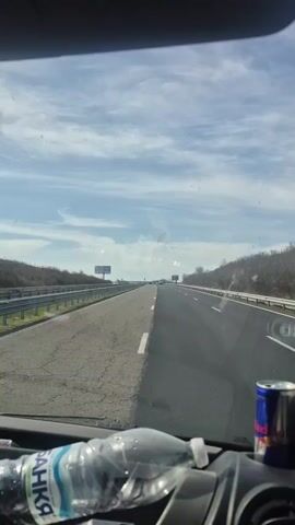 The view from inside a vehicle driving on a highway shows a clear, sunny sky with scattered clouds overhead. The highway has two lanes separated by a dotted white line, with a mix of asphalt textures visible. A gray guardrail lines the side of the road, and sparse trees and foliage can be seen on the outskirts. On the dashboard, a bottle of "BANKIA" water and a can of Red Bull are positioned. In the distance, large billboards are visible on both sides of the highway. Another vehicle can be seen ahead in the distance.